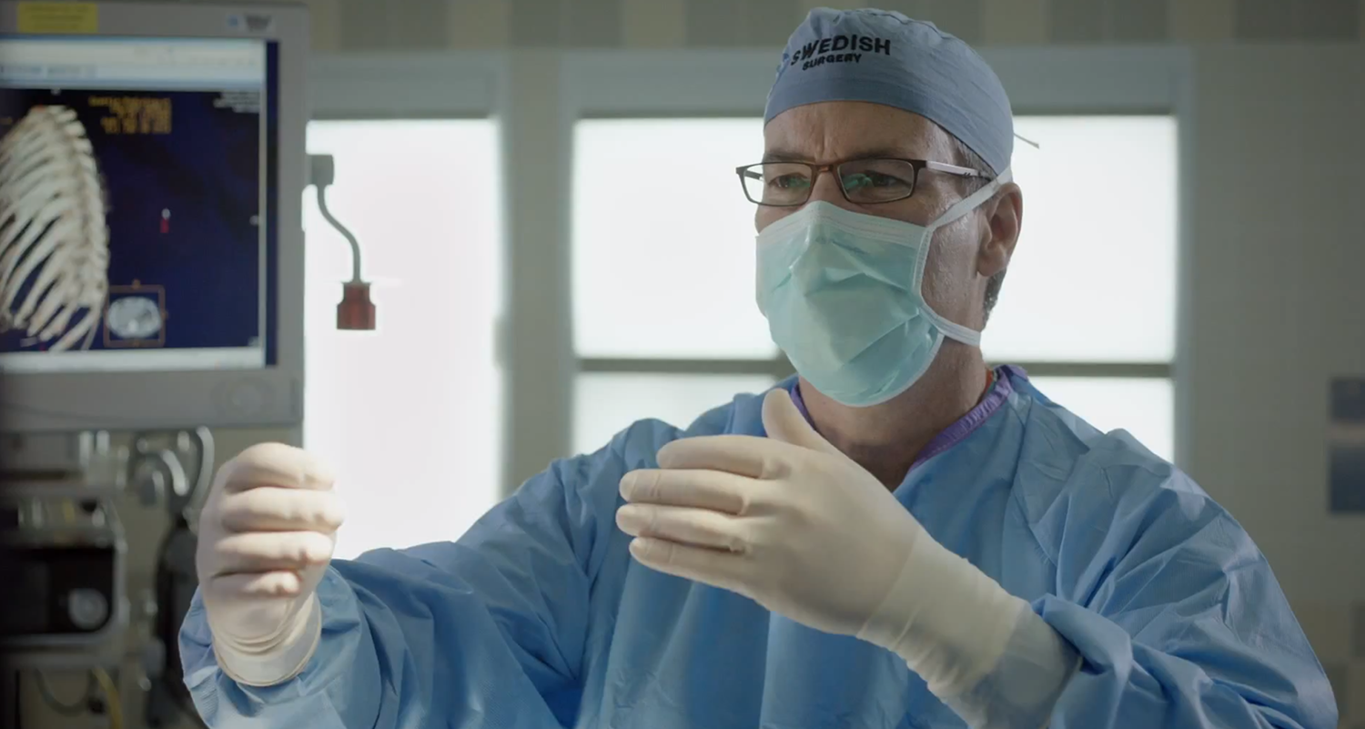 A surgeon in scrubs and mask gesturing with both hands to control imaging on an operating room display.