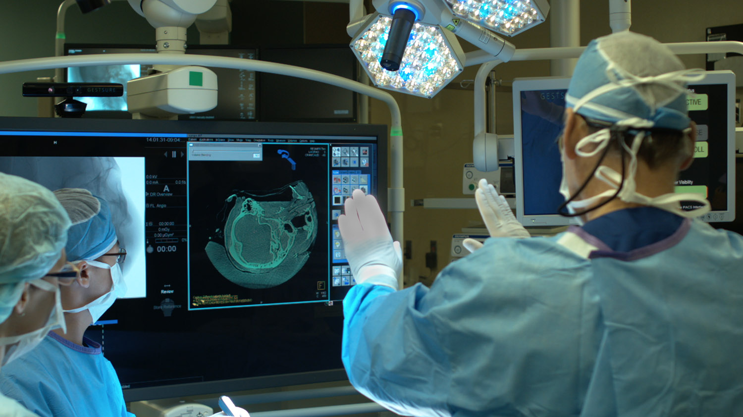 Two surgeons in an operating room reviewing a CT scan on a monitor, one directing it with a raised gloved hand.